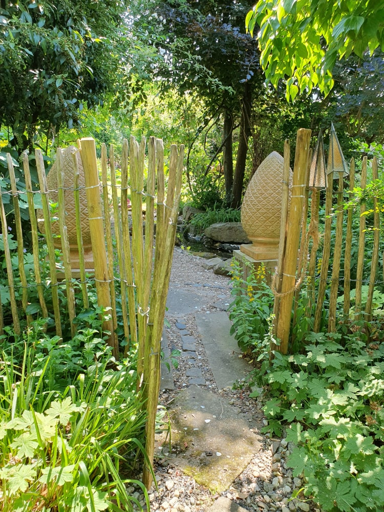 Ein Stück Zaun mit einem Tor trennt den Garten. Es sind zwei große Zapfen aus Ton zu sehen.
