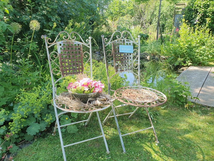Zwei schmiedeeiserne Stühle stehen vor einem Teich auf einer Wiese. Auf einem Stuhl ist eine Schüssel mit Blumen.