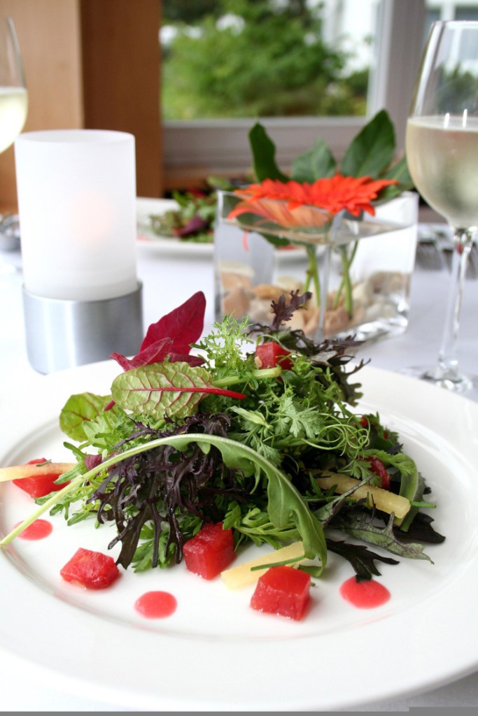 Ein Wildkrautsalat auf einem Tisch mit weißer Tischdecke, Blumen, Kerze und einem Glas Wein.